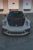 A high-performance sports car is parked in front of a beige industrial building with three white garage doors. The car has a sleek design with a black hood and front grille.