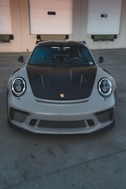 A high-performance sports car is parked in front of a beige industrial building with three white garage doors. The car has a sleek design with a black hood and front grille.
