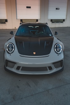 A high-performance sports car is parked in front of a beige industrial building with three white garage doors. The car has a sleek design with a black hood and front grille.