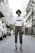 A person stands confidently in an urban street setting, wearing a white t-shirt with black graphics, brown plaid pants, striped socks, black high-top sneakers, and a navy bucket hat. The street is flanked by buildings and power lines, with several parked vehicles visible in the background.
