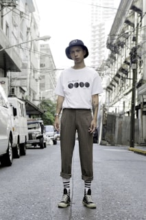 A person stands confidently in an urban street setting, wearing a white t-shirt with black graphics, brown plaid pants, striped socks, black high-top sneakers, and a navy bucket hat. The street is flanked by buildings and power lines, with several parked vehicles visible in the background.