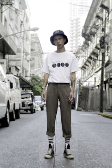 A person stands confidently in an urban street setting, wearing a white t-shirt with black graphics, brown plaid pants, striped socks, black high-top sneakers, and a navy bucket hat. The street is flanked by buildings and power lines, with several parked vehicles visible in the background.