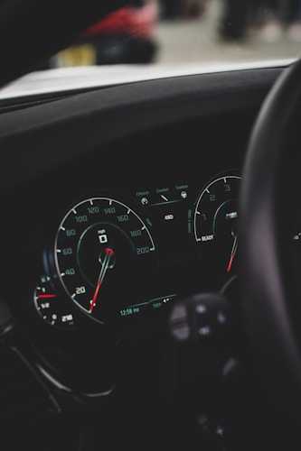 A close-up view of a car dashboard featuring speedometers with marked speeds up to 200 mph. There are indicators for fuel efficiency modes and a display showing the time and odometer reading. The image captures parts of the steering wheel and hints of the exterior environment through reflections.