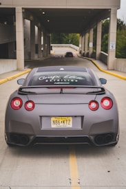 A sleek sports car is parked in the middle of a driveway leading into a well-lit parking garage. The car has a matte finish and visible branding on the rear window. The license plate indicates it is registered in New Jersey.