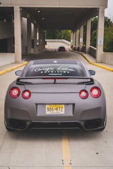 A sleek sports car is parked in the middle of a driveway leading into a well-lit parking garage. The car has a matte finish and visible branding on the rear window. The license plate indicates it is registered in New Jersey.