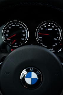 View of a car dashboard showing the speedometer and tachometer, with the BMW logo prominently displayed on the steering wheel. The dials are illuminated with red needles and white numbers, suggesting a sleek and modern design.