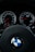 View of a car dashboard showing the speedometer and tachometer, with the BMW logo prominently displayed on the steering wheel. The dials are illuminated with red needles and white numbers, suggesting a sleek and modern design.