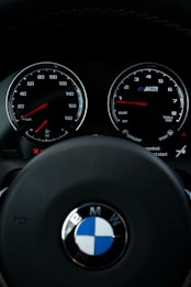 View of a car dashboard showing the speedometer and tachometer, with the BMW logo prominently displayed on the steering wheel. The dials are illuminated with red needles and white numbers, suggesting a sleek and modern design.
