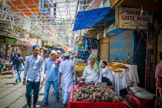 A bustling street market scene featuring a mix of vendors and shoppers. A vendor is prominently visible in the foreground, displaying a variety of goods such as vegetables. People walking by are dressed in traditional attire, and the street is decorated with colorful streamers overhead. Various signs in different languages are visible, indicating a local, vibrant market atmosphere.