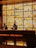 Two men stand at a wooden reception desk in front of a large, illuminated wall featuring a geometric design of vertical orange and yellow stripes. The ambient lighting creates a warm, cozy atmosphere, and a small decorative object is placed on a shelf in the background.