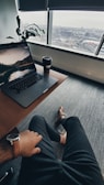 A relaxed workspace setting with an individual sitting at a desk. A laptop displaying a scenic wallpaper and a takeaway coffee cup are on the desk. A large window provides a view of an urban landscape. The person wears a watch and dress shoes, adding a touch of professionalism. A potted plant sits in the corner, adding greenery to the scene.