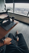 A relaxed workspace setting with an individual sitting at a desk. A laptop displaying a scenic wallpaper and a takeaway coffee cup are on the desk. A large window provides a view of an urban landscape. The person wears a watch and dress shoes, adding a touch of professionalism. A potted plant sits in the corner, adding greenery to the scene.
