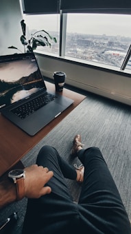 A relaxed workspace setting with an individual sitting at a desk. A laptop displaying a scenic wallpaper and a takeaway coffee cup are on the desk. A large window provides a view of an urban landscape. The person wears a watch and dress shoes, adding a touch of professionalism. A potted plant sits in the corner, adding greenery to the scene.