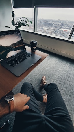 A relaxed workspace setting with an individual sitting at a desk. A laptop displaying a scenic wallpaper and a takeaway coffee cup are on the desk. A large window provides a view of an urban landscape. The person wears a watch and dress shoes, adding a touch of professionalism. A potted plant sits in the corner, adding greenery to the scene.