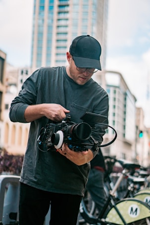A person wearing a cap and glasses is operating a professional video camera on a city street, with tall buildings in the background and bicycles nearby.