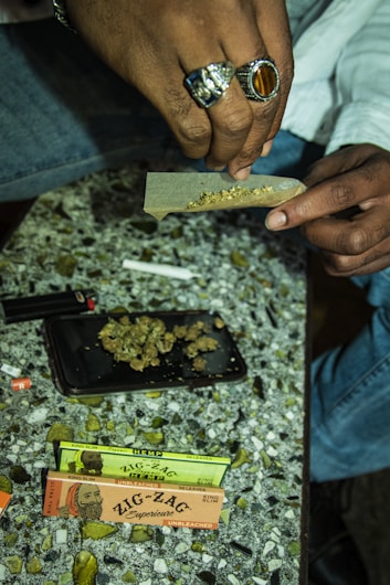 A person is rolling a joint on a speckled surface using a rolling paper. There are two packs of Zig-Zag rolling papers and some dried plant material on a tray. The person is wearing a silver ring with a large stone. A lighter and other small items are also present on the table.