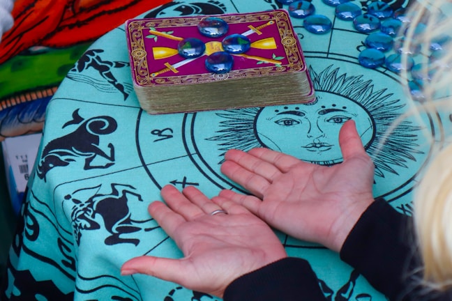 A pair of hands is positioned palms-up on a cloth featuring zodiac signs and a sun-like face. A stack of tarot cards, adorned with mystical symbols, rests on the cloth, surrounded by scattered blue glass beads.