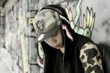 A person wearing a hooded jacket with a shark design on the hood and camouflage patterns on the sleeves is leaning against a graffiti-covered wall. The individual is also wearing a camouflage hat with a sports team logo.