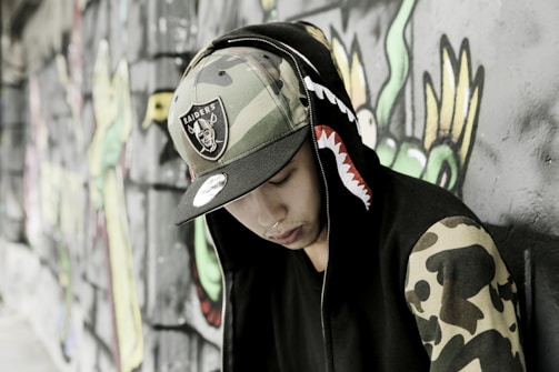 A person wearing a hooded jacket with a shark design on the hood and camouflage patterns on the sleeves is leaning against a graffiti-covered wall. The individual is also wearing a camouflage hat with a sports team logo.