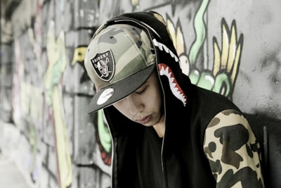 A person wearing a hooded jacket with a shark design on the hood and camouflage patterns on the sleeves is leaning against a graffiti-covered wall. The individual is also wearing a camouflage hat with a sports team logo.