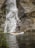 Two people are on a paddleboard in front of a cascading waterfall, surrounded by rocky cliffs and a calm body of water. One person is standing and paddling, while the other is seated. The scene is lush with greenery, and the overall mood is serene and adventurous.