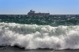 A cargo ship is sailing on the horizon across a vast ocean, with large waves crashing in the foreground under a clear blue sky.