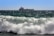 A cargo ship is sailing on the horizon across a vast ocean, with large waves crashing in the foreground under a clear blue sky.