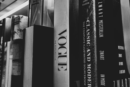 A collection of books is arranged upright on a shelf, with titles visible on their spines. The prominent book is titled 'Vogue: The Editor's Eye', and adjacent to it is 'Classic and Modern: The Complete Illustrated Sourcebook'. The image is in black and white, giving a classic feel to the scene.