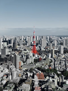 A bustling cityscape with a prominent red and white tower standing out among numerous high-rise buildings. The landscape extends to the horizon under a clear sky, showcasing an urban environment with a mix of modern and traditional architecture.