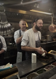 Two chefs are in a busy commercial kitchen, both wearing striped aprons and white shirts. One chef is smiling and holding a block of cheese, while the other adjusts his apron from behind. There are various kitchen utensils and pots hanging above, along with ingredients organized in containers on the stainless steel counter.