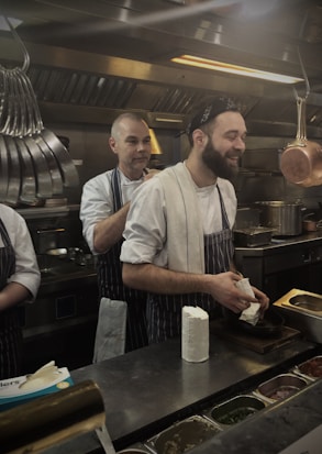 Two chefs are in a busy commercial kitchen, both wearing striped aprons and white shirts. One chef is smiling and holding a block of cheese, while the other adjusts his apron from behind. There are various kitchen utensils and pots hanging above, along with ingredients organized in containers on the stainless steel counter.