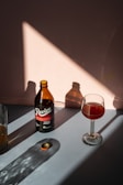 A brown bottle labeled 'Revive Kombucha Hibiscus Refresher' stands upright on a surface, casting a shadow towards the left. Next to it, a wine glass contains a reddish liquid, also casting a shadow. A bottle cap lies nearby, illuminated by a triangular patch of sunlight. The background features warm, muted tones and soft shadows.