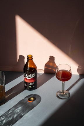 A brown bottle labeled 'Revive Kombucha Hibiscus Refresher' stands upright on a surface, casting a shadow towards the left. Next to it, a wine glass contains a reddish liquid, also casting a shadow. A bottle cap lies nearby, illuminated by a triangular patch of sunlight. The background features warm, muted tones and soft shadows.