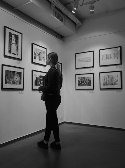 A person stands in an art gallery observing framed photographs mounted on the walls. The space features overhead lighting with a minimalist and modern interior design. The photographs appear to be black and white, enhancing the calm and contemplative atmosphere.