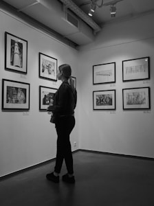 A person stands in an art gallery observing framed photographs mounted on the walls. The space features overhead lighting with a minimalist and modern interior design. The photographs appear to be black and white, enhancing the calm and contemplative atmosphere.