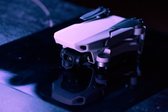 A small drone with a sleek, compact design is resting on a reflective surface under low light, casting a faint reflection. The image emphasizes the drone's aerodynamic shape and minimalistic form.