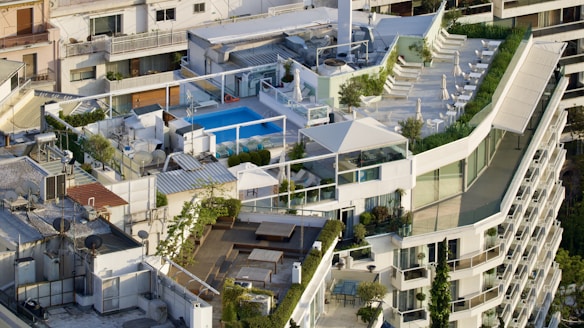 An aerial view of a rooftop complex featuring a swimming pool, several seating areas, and lush greenery. The design incorporates modern architecture with white and glass elements, and there is a spacious terrace with lounge chairs and umbrellas. Various residential buildings surround the rooftop space.