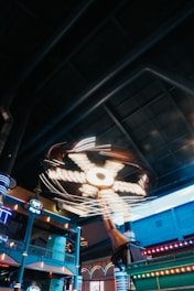An indoor amusement ride features bright, spinning lights and a large structure with moving parts. The ride is in a spacious area with colorful pillars and architectural elements that suggest a fun, entertaining environment.