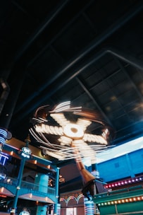 An indoor amusement ride features bright, spinning lights and a large structure with moving parts. The ride is in a spacious area with colorful pillars and architectural elements that suggest a fun, entertaining environment.