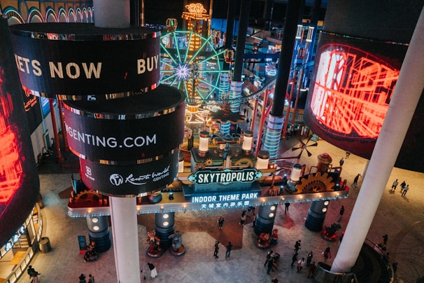 An indoor theme park with vibrant, colorful lights and signage. The neon lights are abundant, featuring circular screens displaying advertisements. A large sign reads 'Skytropolis Indoor Theme Park' surrounded by various attractions including a Ferris wheel.