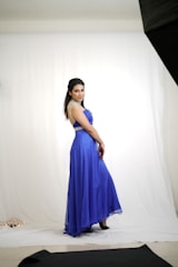 A person in a striking blue gown poses gracefully in a studio setting. The gown features elegant embellishments on the shoulders and waist, enhancing its sophisticated look. The clean white backdrop and soft lighting add to the image's focus on the individual and their attire.