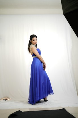 A person in a striking blue gown poses gracefully in a studio setting. The gown features elegant embellishments on the shoulders and waist, enhancing its sophisticated look. The clean white backdrop and soft lighting add to the image's focus on the individual and their attire.