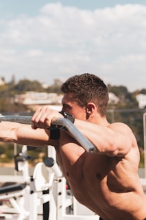 A fit, shirtless person performing an exercise on an outdoor gym machine, with a focus on their muscular physique. The background features a clear sky and blurred scenery with buildings and trees.