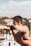 A fit, shirtless person performing an exercise on an outdoor gym machine, with a focus on their muscular physique. The background features a clear sky and blurred scenery with buildings and trees.