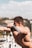 A fit, shirtless person performing an exercise on an outdoor gym machine, with a focus on their muscular physique. The background features a clear sky and blurred scenery with buildings and trees.