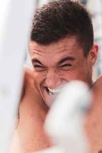 A close-up of a person with a tanned complexion, their facial expression intense and focused, possibly mid-activity. The person has short, dark hair and is possibly outdoors based on lighting.