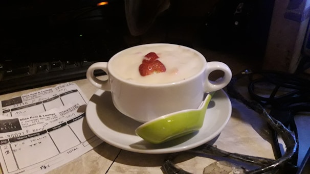 A white ceramic cup filled with creamy yogurt and topped with sliced strawberries beside a green spoon. The cup is on a white saucer. Nearby on the table, there appears to be a receipt or a bill. The background includes some electronic equipment and cables.