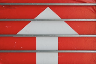 A large white upward arrow is set against a vibrant red background. Four parallel gray metal rods run horizontally across the scene, intersecting the arrow.