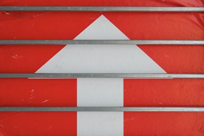 A large white upward arrow is set against a vibrant red background. Four parallel gray metal rods run horizontally across the scene, intersecting the arrow.