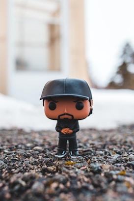 A small vinyl figure is positioned on a rough, gravel-like surface. The figure has a stylized head with a beard and is wearing a black cap and black clothing, including a jacket and pants. The background is blurred, consisting of soft colors suggesting an outdoor environment with some blurred trees and structures.